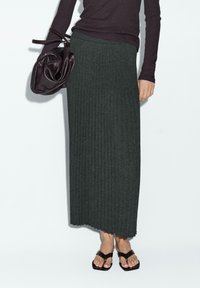 Grauer, gerippter Maxi-Rock mit strukturiertem Finish, kombiniert mit einem dunklen Oberteil und einer geschlossenen Ledertasche. Sandalen runden den Look ab.