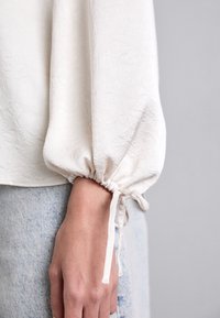 Blusa bianca con texture, maniche arricciate e lacci ai polsini, con una finitura liscia. La blusa è abbinata a un tessuto di denim.