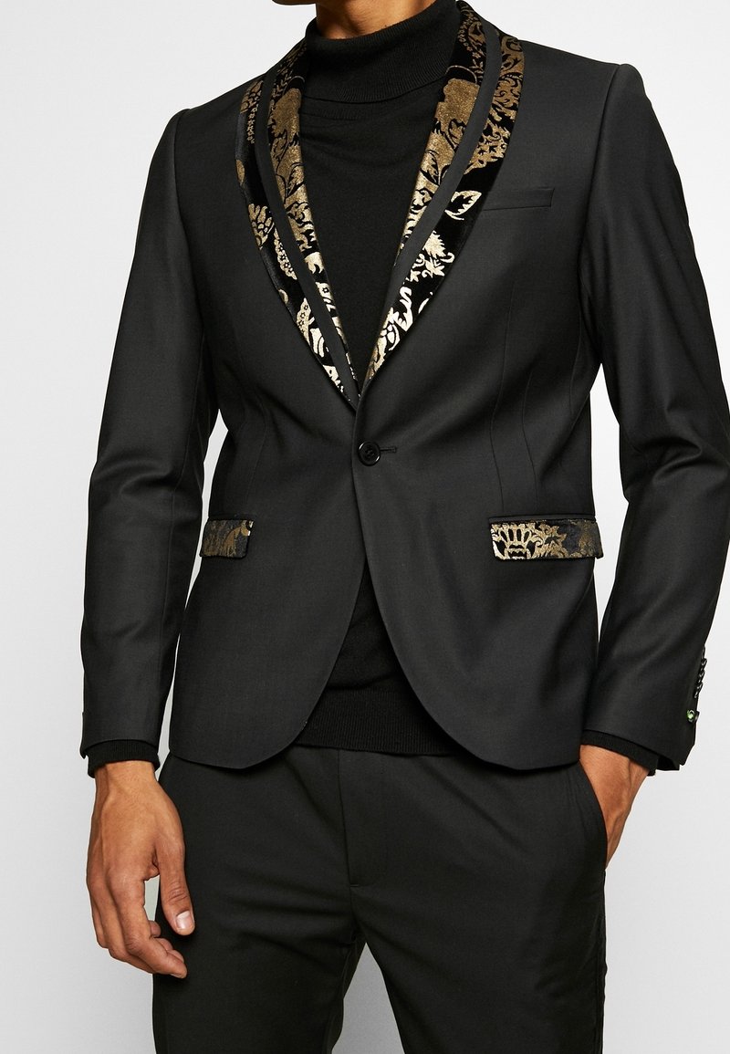 Schwarzer Blazer mit einem gemusterten goldenen und schwarzen Schalkragen, glatter Textur, einreihiger Knopfverschluss und akzentuierten Taschen. Über einem schwarzen Rollkragenpullover getragen.