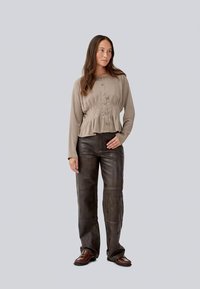 Blusa larga de color marrón claro, con mangas largas y botón en la parte delantera, con una cintura plisada, combinada con pantalones de cuero marrón oscuro y zapatos marrones sobre un fondo claro.