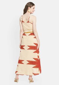 Robe maxi en tissu beige clair avec des motifs géométriques rouges. Elle possède des bretelles volantes, un buste ajusté et une jupe fluide.