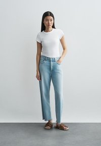 Λευκό ριγωτό μπλουζάκι συνδυασμένο με ανοιχτό μπλε τζιν παντελόνι κατευθείαν γραμμής. Το τζιν έχει φθαρμένο τελείωμα, και το μοντέλο φοράει καφέ σανδάλια.