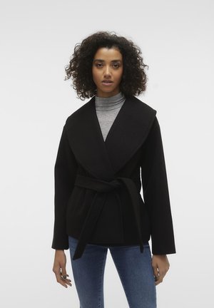 Veste noire à enrouler avec un large col, des manches longues et une ceinture à la taille. Associée à un pull col roulé gris et un jean bleu. Texture de tissu lisse.
