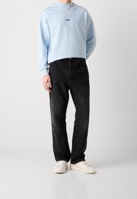 Felpa azzurra chiara con colletto rotondo e piccolo logo, abbinata a jeans neri e sneakers bianche, esposta su uno sfondo neutro.