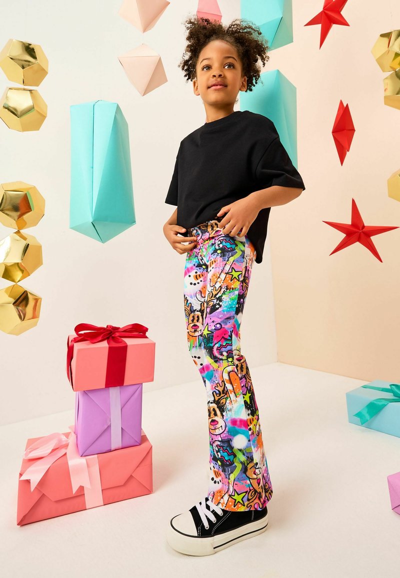 Lapsi, jolla on värikkäät poroprinttipöksyt ja musta paita, seisoo pinottujen lahjakoristen vieressä, joissa on riippuvia geometristen ja tähden muotoisia koristeita.