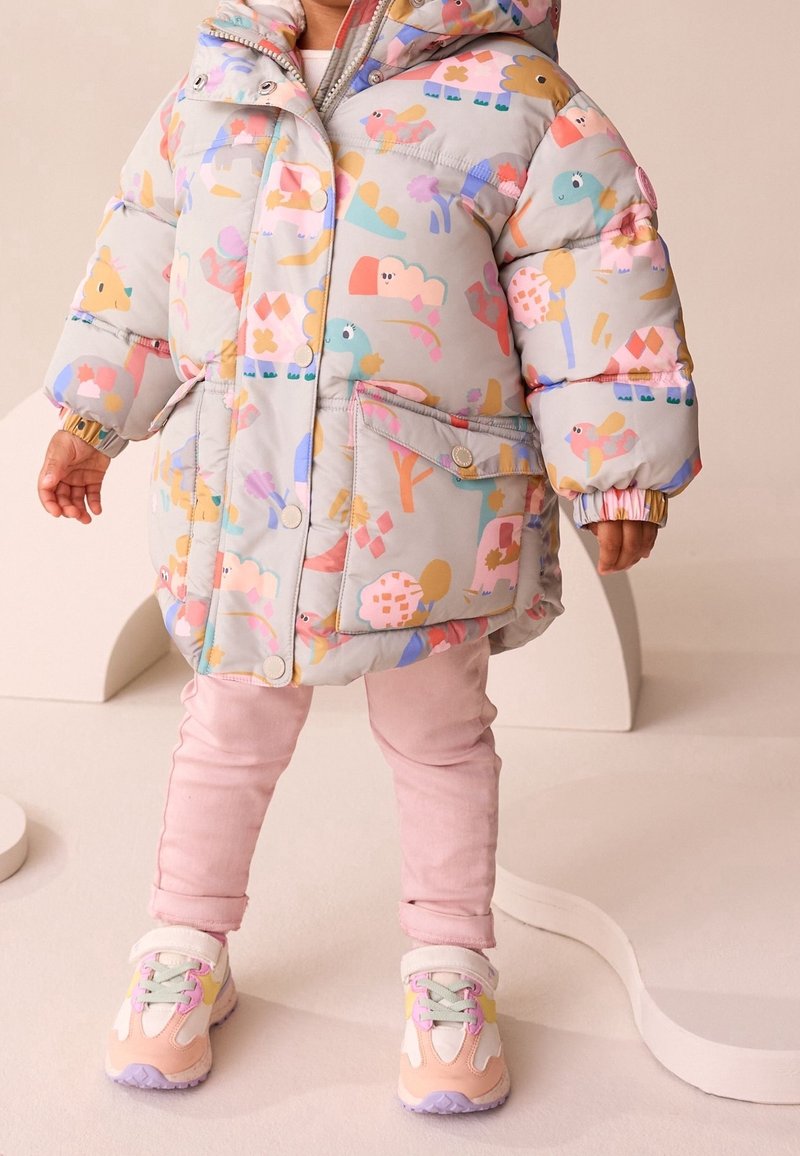 Tout-petit portant une doudoune gris clair avec des motifs d’animaux colorés, un pantalon rose clair et des baskets pastel, debout sur un sol beige.
