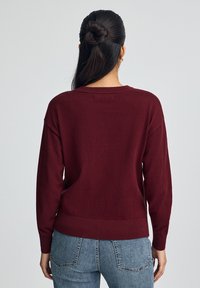 Burgunderfarbener Strickpullover mit lässigem Schnitt, überschnittenen Schultern, geripptem Saum und Bündchen. Getragen mit hellblauen Jeans, die ein lässiges Design zeigen.