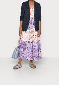 Femme portant une robe florale violette et blanche avec un blazer bleu marine, portant un sac à main bleu clair et des sandales grises.