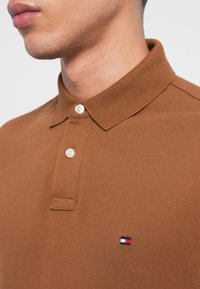 Καφέ πόλο μπλουζάκι με υφή, διαθέτοντας γιακά και μπροστινό μέρος με δύο κουμπιά, διακοσμημένο με ένα μικρό λογότυπο σε κόκκινο, λευκό και μπλε χρώμα.