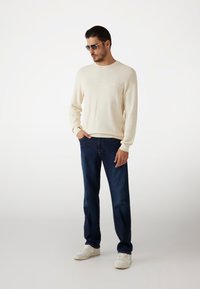 Sweatshirt crème, jean bleu foncé et baskets blanches. Le modèle porte des lunettes de soleil. Tissu doux, design décontracté, coupe relaxe.
