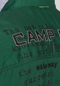 Chemise en tissu vert avec un grand texte brodé en noir indiquant « CAMP » et des mots imprimés en différentes tailles et polices.