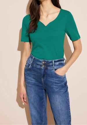 Frau trägt ein eng anliegendes grünes Kurzarmoberteil mit V-Ausschnitt und hoch taillierte blaue Jeans mit doppelten Knöpfen.