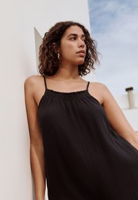 Top nero senza maniche con tessuto strutturato. Presenta una scollatura arricciata e spalline sottili. Lo sfondo è un muro neutro sotto un cielo azzurro.