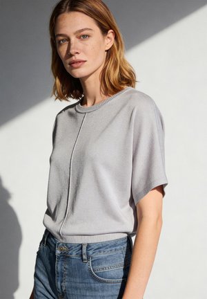 Vrouw met lichtbruin haar tot op de schouders, die een lichtgrijze korte mouwen trui en blauwe spijkerbroek draagt, staand tegen een lichte achtergrond.