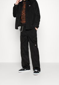 Camicia e pantaloni in velluto a coste neri, con maglione marrone a righe tigre. Scarpe da ginnastica nere con suole bianche, accessoriate con una catenina d'argento.