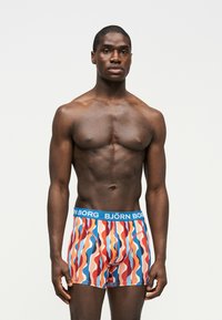 Homme musclé debout devant un fond uni, portant un boxer coloré à motif ondulé avec une ceinture bleue portant l'inscription "BJÖRN BORG."
