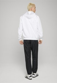 Chaqueta blanca con capucha y puños elásticos, combinada con pantalones negros y zapatillas deportivas blancas con rayas negras. Tejido liso y ligero.