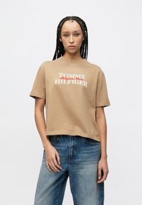 Καφέ κοντομάνικο T-shirt με κοντά μανίκια, με λευκή και κόκκινη επιγραφή "TOMMY HILFIGER" στο μπροστινό μέρος, κατασ�κευασμένο από απαλό βαμβακερό υλικό.