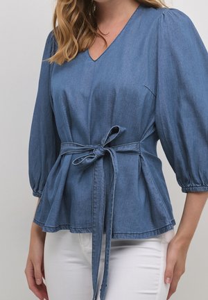 Nainen, joka pukeutunut siniseen farkkujen V-kaula-aukkoinen puseroon, jossa on 3/4-puffihihat ja sidottu vyötärönauha, yhdistettynä valkoisiin housuihin.