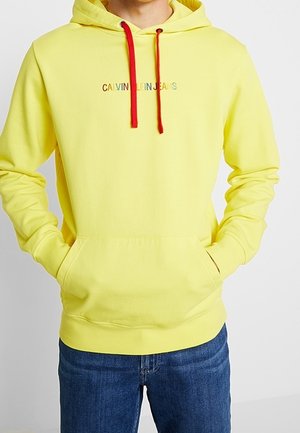 Personne portant un sweat à capuche jaune vif avec des cordons rouges et le logo « Calvin Klein Jeans » en texte multicolore, associé à un jean bleu.