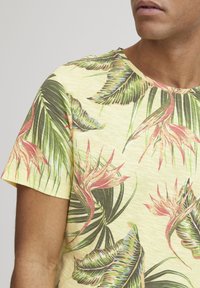 Camiseta de manga corta y cuello redondo en amarillo claro con un patrón de hojas y flores tropicales en verde, rosa y azul. Tela ligera y texturizada.