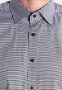 Schwarz-weißer gemusterter Hemd mit Kragen und schwarzen Knöpfen. Der Stoff ist strukturiert mit einem feinen, karierten Design.