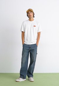 Hvitt bomull T-skjorte med liten rød logo, blå denim jeans, beige caps, og grå sneakers med hvite detaljer. Minimalistisk design, avslappet passform.