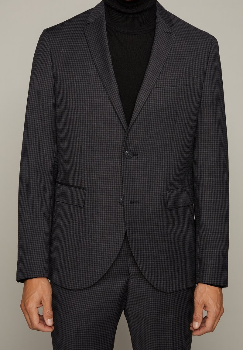 Mörkt rutigt blazer med en slät yta, enkel knappknäppning och två framfickor, bärs över en svart polotröja.