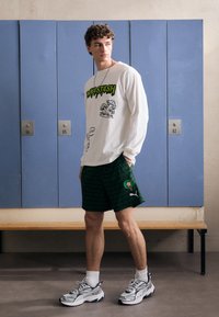 Langærmet hvid t-shirt med grøn grafisk tekst og illustrationer, parret med grønne mønstrede shorts med et logo. Hvide sokker og sneakers.