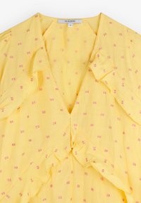 Blouse jaune avec petits motifs carrés roses, encolure en V et bordure froncée le long de l'encolure et de l'ouverture devant, taille moyenne.