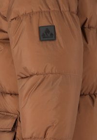 Brun vadderad jacka med ett slätt, strukturerat tyg. Har en svart logotyp-patch på ärmen och en framficka.