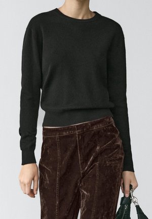 Personne portant un pull noir uni à manches longues et un pantalon en velours marron foncé, tenant un sac à main vert à son côté, sur un fond clair.