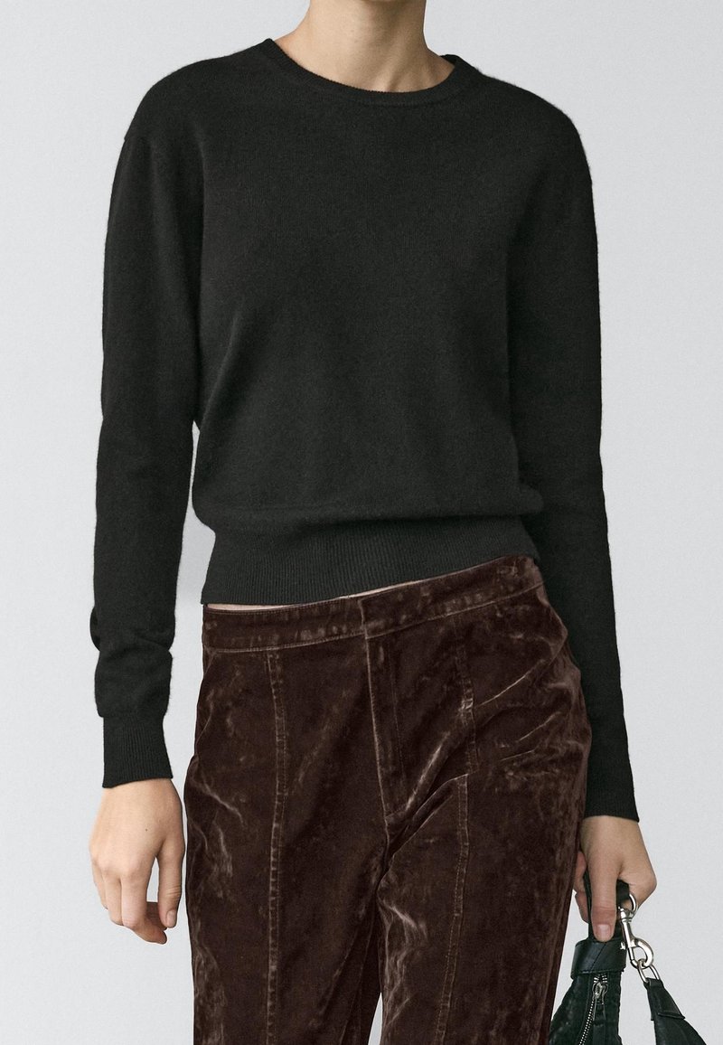 Personne portant un pull noir uni à manches longues et un pantalon en velours marron foncé, tenant un sac à main vert à son côté, sur un fond clair.