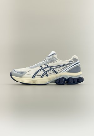 Zapatilla de correr blanca y azul marino con parte superior de malla, suela acolchada y diseño ondulado en el talón sobre un fondo beige liso.