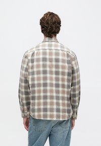 Muž s vlnitými hnedými vlasmi, ktorý má na sebe béžovú a šedú kockovanú košeľu s dlhým rukávom a svetlomodré džínsy, otočený chrbtom na jednoduchom bielom pozadí.