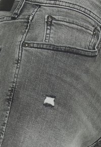 Vaqueros de mezclilla negros con un patrón desgastado, que presentan un gran agujero cuadrado. Los detalles incluyen bolsillos traseros y herrajes metálicos oscuros.