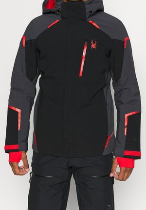 Męska czarno-szara zimowa kurtka z czerwonymi akcentami, kieszenią na piersi na zamek, wysokim kołnierzem i regulowanymi paskami na nadgarstkach, noszona z czarnymi spodniami.