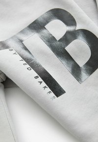 Sudadera gris con una gran letra gráfica en negro "B" y el texto "by Ted Baker", que refleja una textura suave y un patrón sutil.