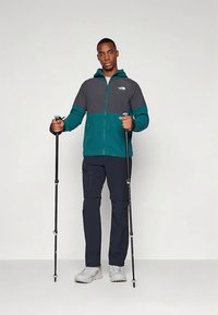 Mees, kes kannab türkiissinist ja halli jope, musti pükse ja valgeid kingi, hoiab musti matkakeppe ning seisab lihtsa tausta ees.