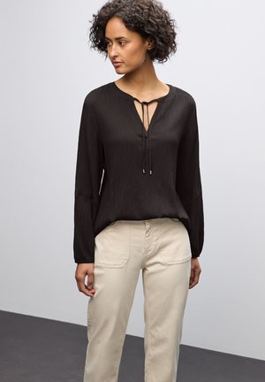Blouse noire à manches longues avec un décolleté en V et un détail à nouer à l'avant ; tissu texturé ; associée à un pantalon beige clair avec poches.