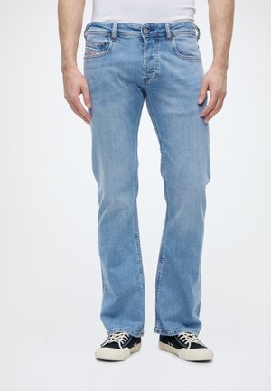Homme portant un jean bleu clair droit, un t-shirt blanc et des baskets noires avec des lacets blancs, debout sur fond blanc.