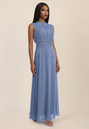 Frau, die ein ärmelloses Kleid mit Spitzenoberteil in Periwinkle-Blau mit gewellten Kanten und einem fließenden Chiffonrock trägt, vor einem beigen Hintergrund stehend.