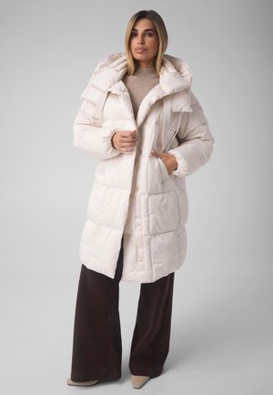 Mujer que lleva un abrigo largo blanco acolchado con un gran cuello, jersey de cuello alto beige, pantalones anchos oscuros y zapatos beige puntiagudos.