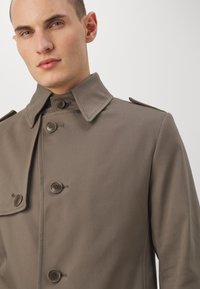Trench coat khaki con colletto strutturato, chiusura con bottoni e spalline sulle spalle. Realizzato in tessuto morbido con una vestibilità su misura.
