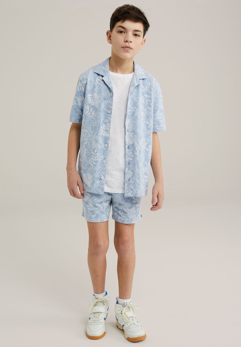 Camicia azzurra a maniche corte e pantaloni corti abbinati con un motivo di felce, abbinati a una maglietta bianca e scarpe alte.