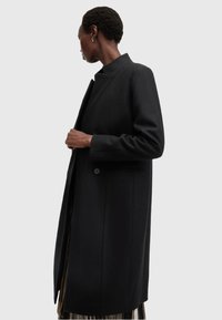 Cappotto in lana nera con colletto alto, silhouette lunga e chiusura con bottoni. Presenta tasche laterali e una superficie liscia.