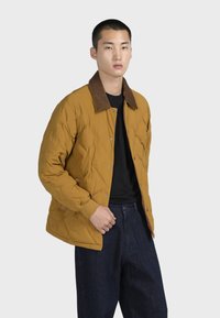 Giacca trapuntata gialla senape con colletto in corduroy marrone, chiusura a scatto anteriore e polsini a costine, abbinata a jeans larghi di denim scuro.