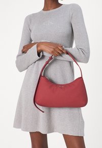 Donna che indossa un vestito a maglia grigio chiaro, mentre tiene in mano una piccola borsa rossa con tracolla corta e chiusura con zip.
