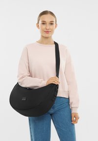 Czarna torba na ramię wykonana z wytrzymałego materiału, o zaokrąglonym kształcie, z regulowanym paskiem i małą łatą z logo z przodu.