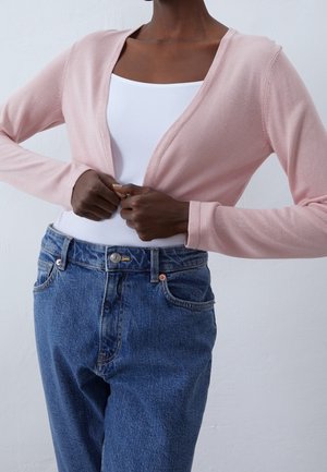 Personne portant un cardigan rose clair par-dessus un haut blanc et un jean bleu taille haute, en train de fermer le cardigan à l'avant.
